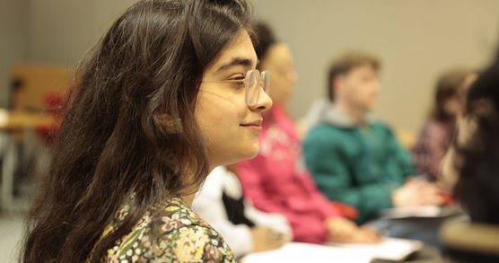 A student wearing glasses, with long brown hair, looking into the distance and smiling. There are other students in the background in a classroom-style environment.