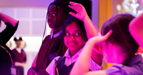 A group of school students in uniform, the child in the centre of the image has raised her hand to answer a question, there is a pink tint to the photo