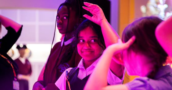 A group of school students in uniform, the child in the centre of the image has raised her hand to answer a question, there is a pink tint to the photo