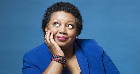 A woman with short black hair, resting her chin on her hand and gazing upwards. She's wearing pink lipstick, a blue suit jacket and colourful bracelets.