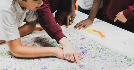 School age students leaning over a map, two of them are reaching into the middle of the map to point out a location