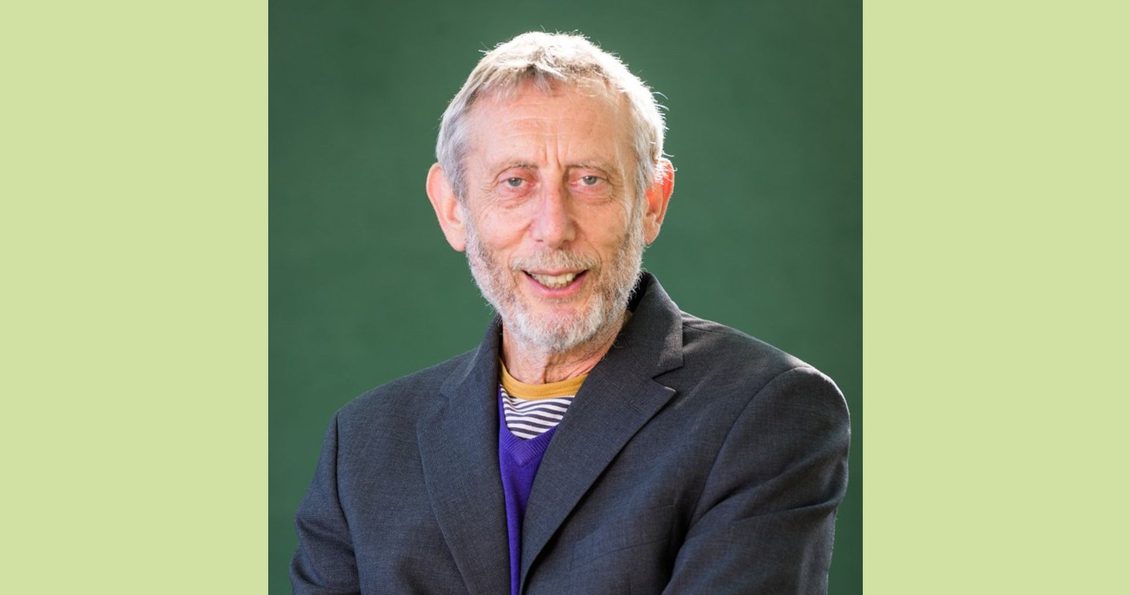 A smiling man with grey hair wearing a bright blue striped top under a grey blazer.