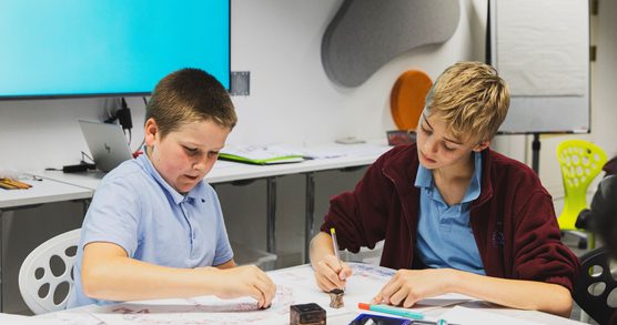 Two young students seated at a desk. They are both writing on a worksheets. They are both wearing blue school polo shirts, the child on the right is also wearing a maroon zip up fleece