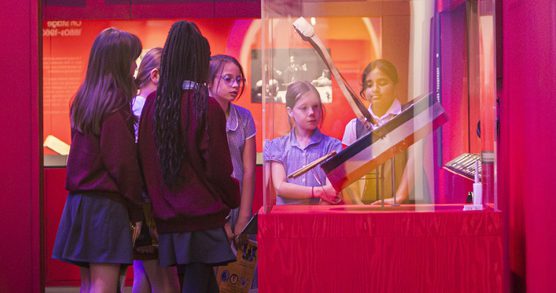A group of 6 schoolgirls of varying heights but all dressed in school uniform have gathered around an exhibition display case to look inside of it.