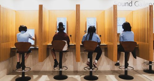 A line of four wooden booths with screen inside the booth. A school age child wearing school uniform is sat with their back to camera on a high stool at each of the booths. All children a wearing headphones.