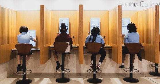 A line of four wooden booths with screen inside the booth. A school age child wearing school uniform is sat with their back to camera on a high stool at each of the booths. All children a wearing headphones.