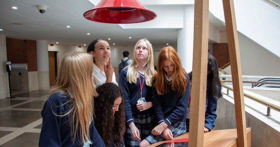 A group of 6 students in blue school jumpers and blue plaid skirts stand around a desk. 3 are looking down at the desk, the other 3 students are looking up at a red funnel which is suspended above them
