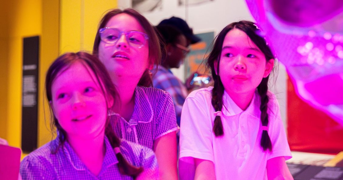 Three uniformed schoolchildren look with interest at something, their faces are lit by a pink light