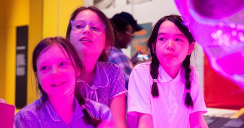 Three uniformed schoolchildren look with interest at something, their faces are lit by a pink light