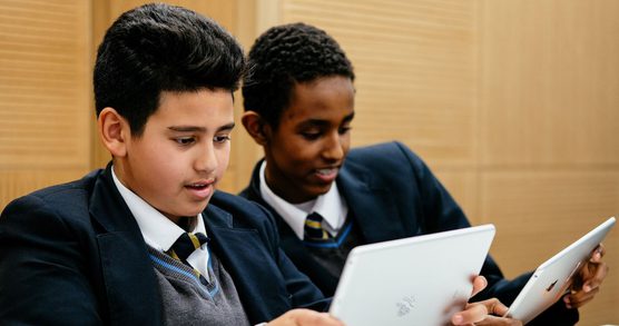 Two schoolboys wearing shcool uniform and holding iPads stand together. They are both looking at their screens, their expressions are amused and interested