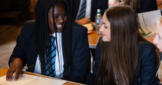 Two students in school uniform sat at a desk. They are looking at each other and smiling, the student on the left has placed a hand on a piece of paper on the desk