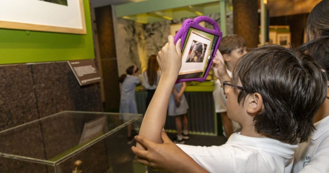 Two primary school students use a tablet to look at and take a photo of an artwork on the wall in a gallery space. Other students can be seen in the background.