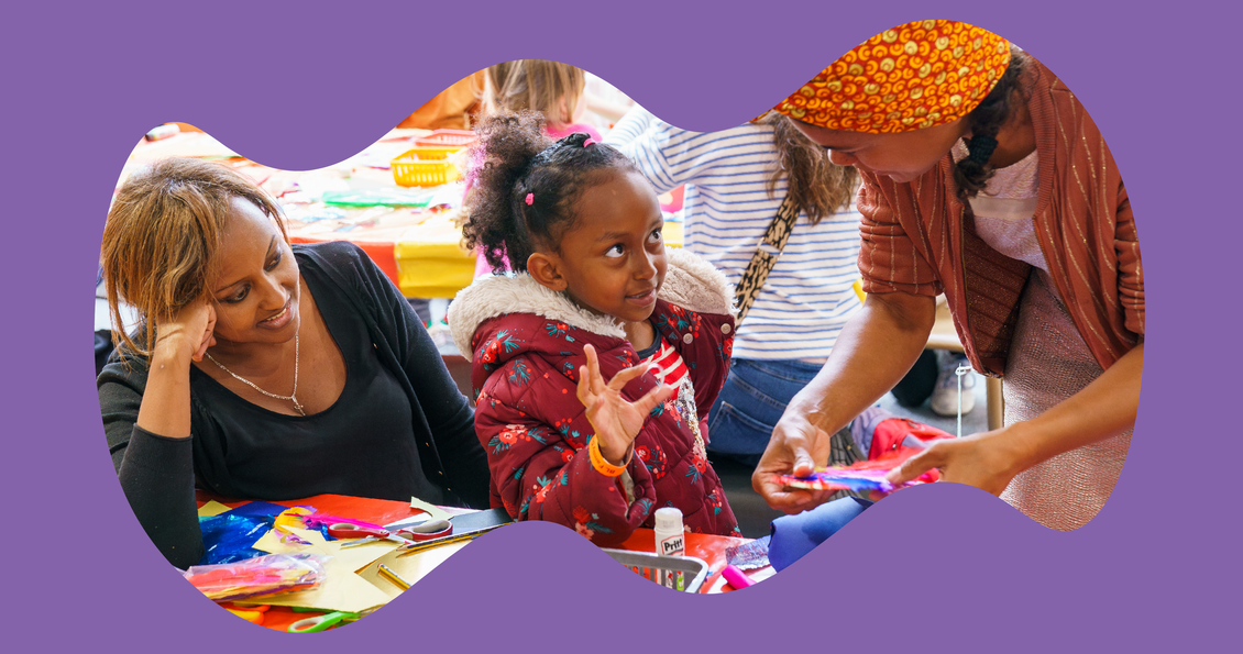 A young girl stands at a craft table with colourful paper and glue sticks on top. She looks up at a woman who is bending down and showing her something in her hands.