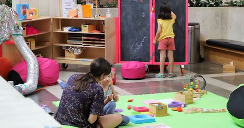 Two children playing, one sits on a mat on the floor with and adult, the other child is drawing on a blackboard