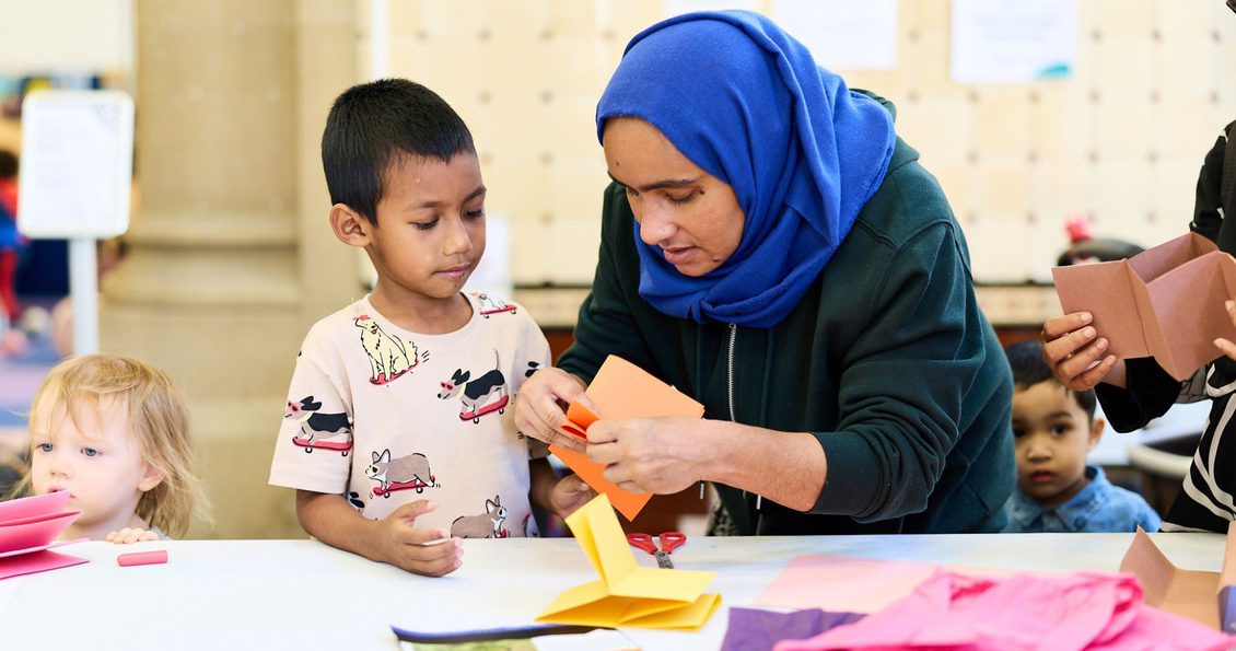 A woman in a blue headscarf crafting with a child. There are colourful pieces of paper on a table and other children in the background.