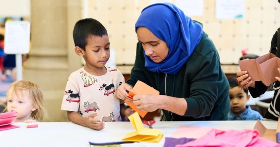 A woman in a blue headscarf crafting with a child. There are colourful pieces of paper on a table and other children in the background.