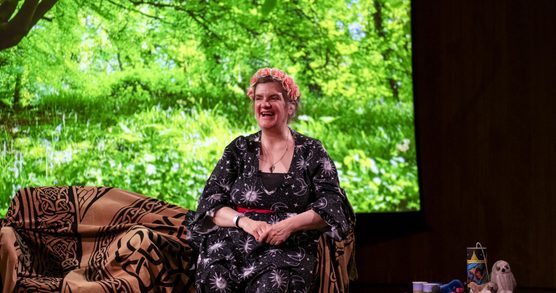 Storyteller Olivia Armstrong sat on a chair with her props on a side table and trees images in the background.