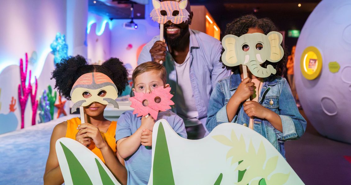 Three children and an adult holding animal masks in front of their face, stood in a colourful room.