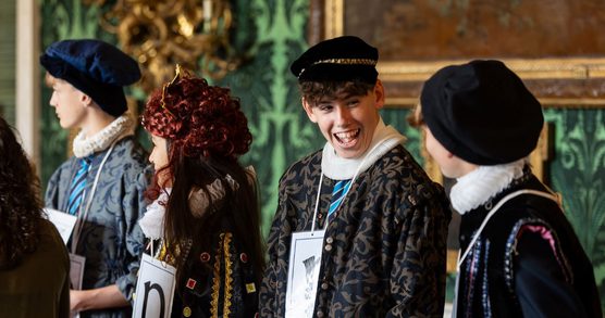 Photograph of children in Tudor costumes included velvet caps and white ruffs, they are standing in an ornately decorated room with green patterned wallpaper and gold picture frames.