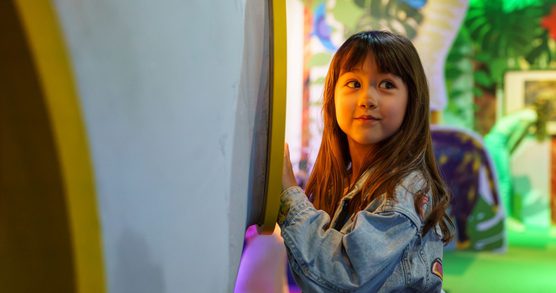 A child glancing past the camera with a smile on her face, stood next to a large blue and yellow structure, with paper trees in the background.