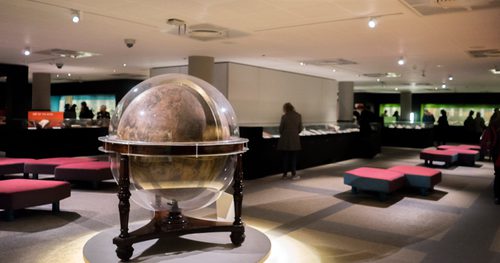 A spot-lit large globe set into a wooden frame standing on a low pedestal in an exhibition gallery